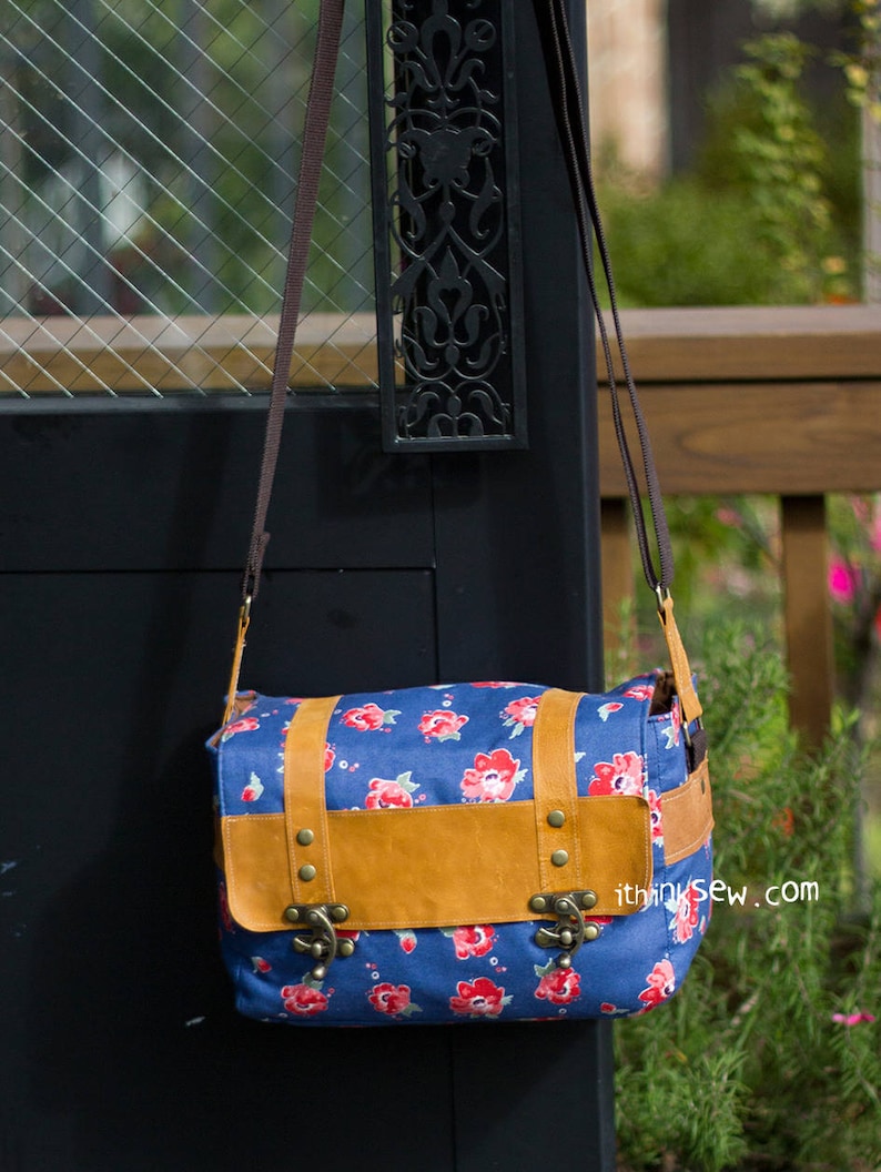 Pu&ograve; includere: Una borsa a tracolla in tela blu con una patta in pelle marrone e fibbie metalliche. La borsa ha una stampa floreale con fiori rossi e dettagli bianchi. La borsa &egrave; appesa a una porta nera con un design in metallo.