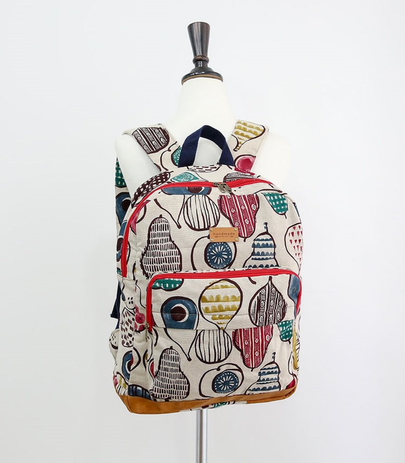 May include: A beige canvas backpack with a red zipper and a brown leather bottom. The backpack is decorated with a colorful pear pattern and the word "handmade" on a brown tag.
