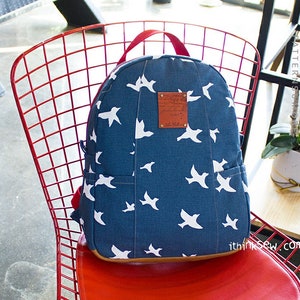 Pode incluir: Uma mochila azul marinho com um padrão de pássaros brancos, alças vermelhas e um remendo de couro castanho. A mochila está em uma cadeira de arame vermelha. A bolsa tem um bolso frontal e uma alça superior.