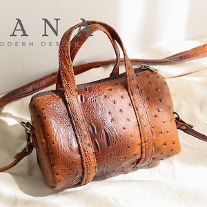 May include: A brown leather barrel bag with a textured ostrich pattern and a crocodile-embossed accent. The bag has a top handle and a detachable shoulder strap. The text "DANA MODERN DESIGN" is at the top.