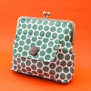 以下が含まれることがあります： 緑と白のドット柄のクラッチバッグ。金属製の留め金と小さなフロントポケットが付いています。ポケットは茶色のボタンで留められています。クラッチバッグはオレンジ色の背景に置かれています。