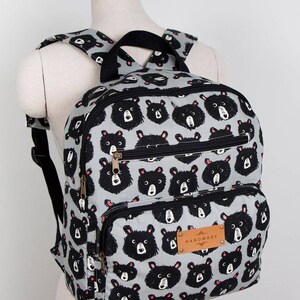 May include: A grey backpack with a black bear print and black straps. The backpack has two zipped pockets and a brown leather tag that says "HANDMADE".