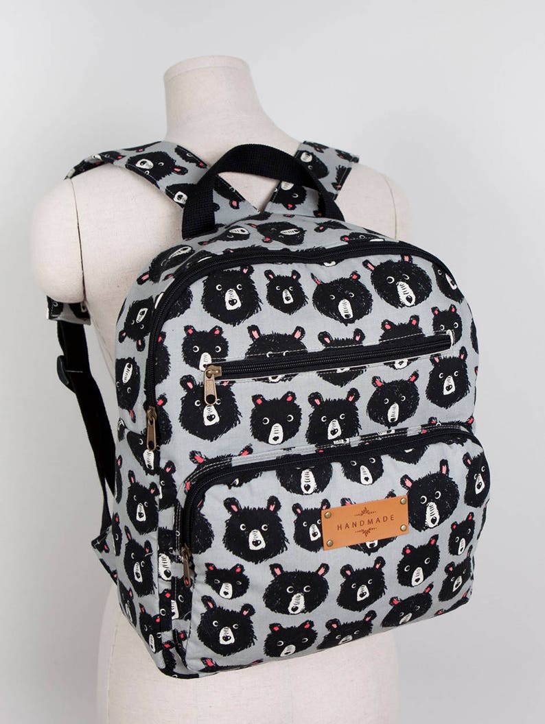 May include: A grey backpack with a black bear print and black straps. The backpack has two zipped pockets and a brown leather tag that says "HANDMADE".