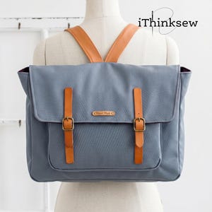 Puede incluir: Una mochila azul pizarra con correas de cuero color canela con hebillas de latón, una solapa frontal y una pequeña etiqueta rectangular con la inscripción Hand Made. La bolsa incluye correas de hombro color canela y se muestra en un maniquí con el texto iThinksew.