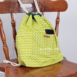 May include: A lime green drawstring backpack featuring a white geometric pattern, a dark green fabric top trim, and thick white rope straps. An external pocket contains a black smartphone and displays a small brown tag labelled HAND MADE.