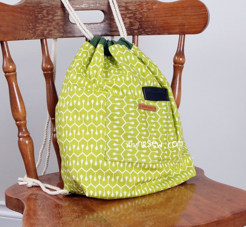 May include: A lime green drawstring backpack featuring a white geometric pattern, a dark green fabric top trim, and thick white rope straps. An external pocket contains a black smartphone and displays a small brown tag labelled HAND MADE.