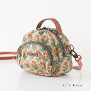Könnte beinhalten: Kleine, runde Handtasche mit einem Blumenmuster in Orange und Grün auf beigem Hintergrund. Sie hat einen braunen Ledergriff und einen abnehmbaren Riemen. Die Tasche hat einen Reißverschluss und das Wort "HANDMADE" ist sichtbar.