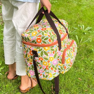 May include: A floral cooler bag with brown straps and trim. The bag features a colourful design of flowers and leaves in shades of pink, orange, yellow, and blue on a white background. The bag has a zipped top and a side pocket.
