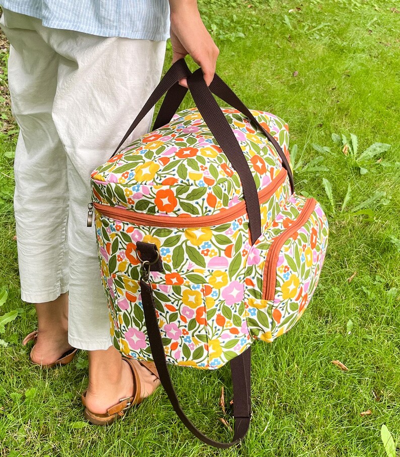 May include: A floral cooler bag with brown straps and trim. The bag features a colourful design of flowers and leaves in shades of pink, orange, yellow, and blue on a white background. The bag has a zipped top and a side pocket.
