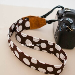 Pu&ograve; includere: Una tracolla per fotocamera a pois marroni e bianchi con una tracolla regolabile in pelle marrone. La tracolla ha il testo "thinksew.com" stampato su di essa.