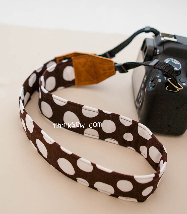 Pu&ograve; includere: Una tracolla per fotocamera a pois marroni e bianchi con una tracolla regolabile in pelle marrone. La tracolla ha il testo "thinksew.com" stampato su di essa.