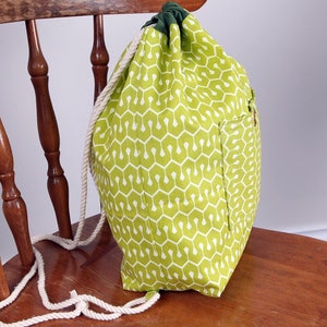 May include: A drawstring backpack featuring a lime green fabric with a repeating white geometric pattern. The bag has a dark green top closure, a side pocket, and thick white twisted rope straps that function as both the closure and shoulder straps.