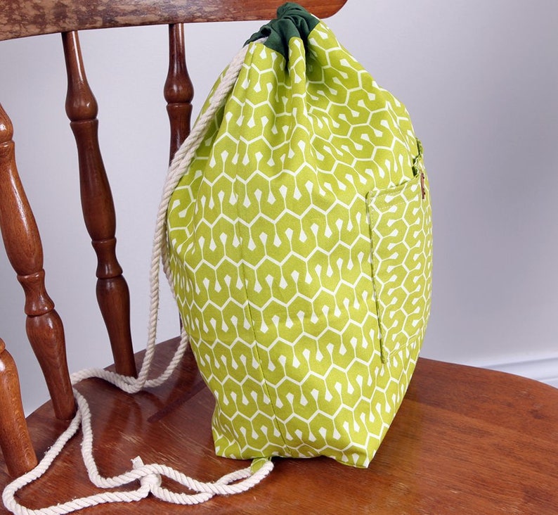 May include: A drawstring backpack featuring a lime green fabric with a repeating white geometric pattern. The bag has a dark green top closure, a side pocket, and thick white twisted rope straps that function as both the closure and shoulder straps.