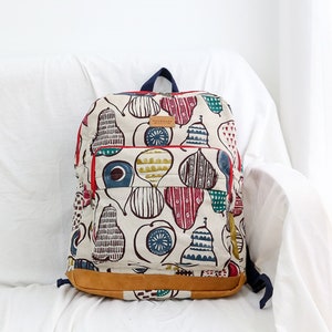 May include: A white canvas backpack with a red zipper and brown leather trim. The backpack is decorated with a colorful pattern of pears and other geometric shapes. The text "handmade" is printed on a brown leather tag.