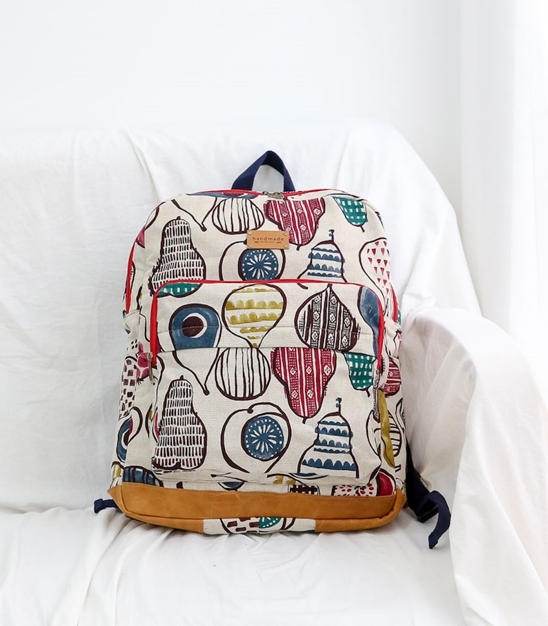 May include: A white canvas backpack with a red zipper and brown leather trim. The backpack is decorated with a colorful pattern of pears and other geometric shapes. The text "handmade" is printed on a brown leather tag.