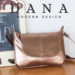 May include: A metallic rose gold colored leather shoulder bag with a flap closure and a long adjustable strap. The bag is sitting on a wooden surface. The text "DANA MODERN DESIGN" is visible in the background.