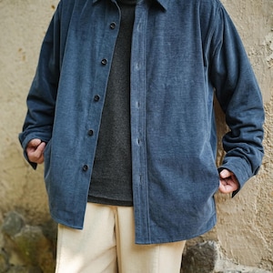 Op de afbeelding: Een blauw corduroy overhemdjack met zwarte knopen. De jas wordt gedragen over een grijze coltrui en gecombineerd met crèmekleurige corduroy broeken. De outfit is casual en comfortabel.