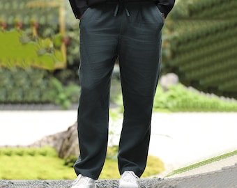 May include: Dark teal linen pants with a drawstring waist and pockets. The pants are paired with a black button-down shirt and white sneakers. The model is standing on a gravel surface with greenery in the background.