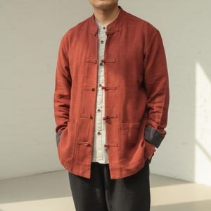 May include: A rust-colored long-sleeve jacket featuring a mandarin collar and a front closure with five fabric-covered toggle buttons. The jacket includes two patch pockets at the waist and dark gray cuffs visible at the sleeves, worn over a light beige shirt.