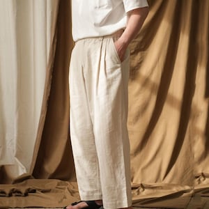 Op de afbeelding: Een persoon draagt een wit T-shirt en een lichtbeige geplooide broek. De broek heeft een relaxte pasvorm en wordt gecombineerd met zwarte sandalen. De achtergrond bestaat uit stoffen draperieën in neutrale tinten.