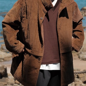 May include: A brown corduroy jacket with a wide collar and button closure. The jacket is layered over a brown sweater and a white t-shirt. The person is wearing dark gray pants.