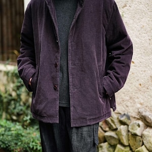May include: A dark purple corduroy jacket with a button-up front and a high collar. The jacket is worn over a gray turtleneck sweater. The person is also wearing dark gray pants. The outfit is casual and stylish.