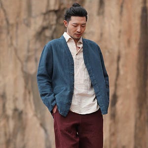 Puede incluir: Una chaqueta azul con cuello en V, usada sobre una camisa de color claro, combinada con pantalones burdeos. La chaqueta tiene botones. El atuendo es informal y parece estar hecho de lino o una tela similar.