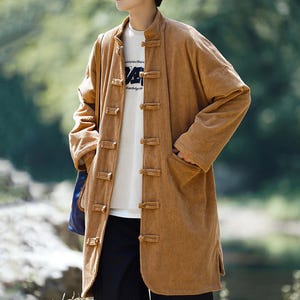 Puede incluir: Un abrigo largo de pana color marrón claro con cuello mandarín y cierres de rana. El abrigo tiene mangas largas y bolsillos. Debajo se lleva una camiseta blanca. Una prenda de vestir exterior elegante.