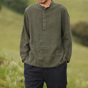 May include: Olive green long-sleeve shirt with a mandarin collar and buttoned placket. The shirt is paired with black pants. The shirt has a relaxed fit and is made of a textured fabric. The background is a blurred green field.