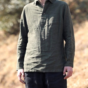 May include: An olive green long-sleeve shirt with a collar and a partial button-down placket featuring a single visible button. The shirt has a textured fabric appearance and is paired with black trousers, creating a simple and functional outfit.