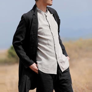 May include: A black linen blazer with a long, flowing cut. The blazer is worn with a light grey button-down shirt and black drawstring trousers. The outfit is suitable for a casual or semi-formal occasion.