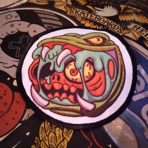 May include: A circular embroidered patch featuring a graphic illustration of a green snake head with a wide-open mouth, sharp white fangs, and a red interior. The snake has a yellow eye with an X-shaped pupil and light blue slime dripping down.
