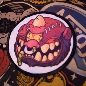 May include: A circular embroidered patch featuring a stylized, cartoonish monster head with a red and purple color scheme. The creature has a large, toothy grin, a single visible eye with an X-shaped pupil, a nose ring, and visible stitches.