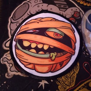 May include: A circular embroidered patch featuring a cartoon mummy character with orange bandages, a single visible red button eye, and a wide mouth with yellow teeth and a small amount of green drool, all outlined in a thick black border.