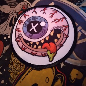 May include: A circular embroidered patch featuring a cartoon eyeball with a purple iris, a white X-shaped pupil, and red blood vessels. The eyeball has a wide, toothy grin with a protruding red tongue dripping with a small amount of green slime.