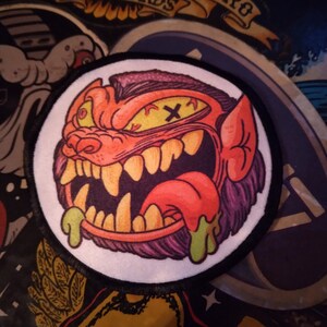 May include: A circular embroidered patch featuring a stylized orange monster head with a purple mohawk, sharp yellow teeth, a protruding tongue, and green slime dripping from its mouth. The monster has one yellow eye and one black X.