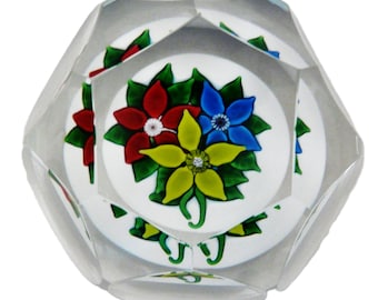 May include: A clear, faceted glass paperweight with a floral design. The design features red, yellow, and blue flowers with green leaves on a white background. The paperweight has a geometric shape.