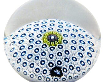 May include: A clear glass paperweight with a white base, featuring a pattern of blue and white circles. The center has a green and purple design. The paperweight has a rounded top and is set against a white background.