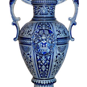Può includere: Un alto vaso in ceramica a due manici con finitura in smalto salino blu e bianco. Il corpo è decorato con complessi motivi in rilievo, tra cui uno stemma centrale raffigurante un castello, un motivo a testa di leone sul collo e figure scolpite.