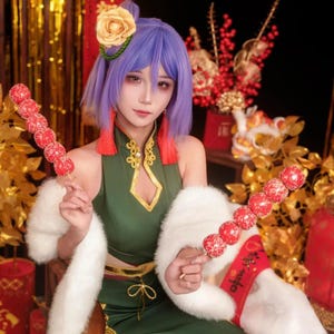 May include: A person in a green dress with gold trim and a white fur stole, holding two skewers of red candied fruit. The person has purple hair and is wearing a flower in their hair. The background includes gold and red decorations.