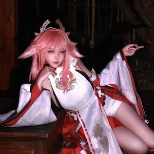 Puede incluir: Una persona con un atuendo de cosplay con cabello rosado y una prenda estilo kimono blanco y rojo. El atuendo incluye un tocado decorativo y una pieza para el pecho con un emblema dorado. El fondo es oscuro.