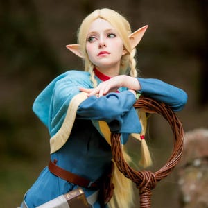 Könnte beinhalten: Eine Person in einem Cosplay-Kostüm mit langen blonden Haaren, spitzen Ohren und einer blauen Tunika mit braunem Gürtel. Die Person lehnt sich an eine braune, kreisförmige Requisite, mit verschwommenem Hintergrund.