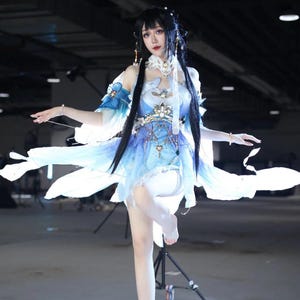 May include: A light blue and white cosplay dress with intricate floral and star-like embellishments. The dress features a layered skirt and flowing sleeves. Long black hair and delicate jewelry complete the look.