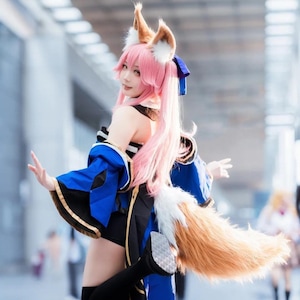 Könnte beinhalten: Ein Cosplay-Outfit mit rosa Perücke, Fuchsohren und einem langen, flauschigen Schwanz. Das Kostüm beinhaltet ein blaues und schwarzes Kleid mit goldenen Verzierungen. Die Person posiert im Freien.