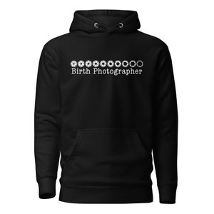 Könnte beinhalten: Schwarzer Kapuzenpullover mit Fronttasche und Kordelzugkapuze. Der weiße Text "Birth Photographer" ist unter einer Reihe weißer Kamera-Blenden-Symbole zentriert. Ein lässiges, bequemes Kleidungsstück.
