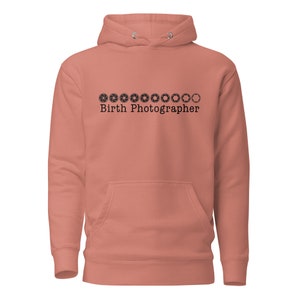 Könnte beinhalten: Ein lachsfarbener Hoodie mit einer schwarzen Grafik von Kamera-Blenden-Symbolen und dem Text "Birth Photographer".