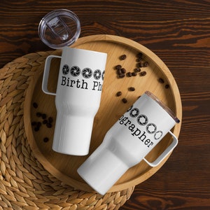Könnte beinhalten: Zwei weiße Edelstahl-Reisebecher mit schwarzem Text und Kameraobjektiv-Grafiken. Der Text auf den Bechern lautet "Birth Photographer" und "Photographer". Die Becher stehen auf einem Holztablett mit Kaffeebohnen, die um sie herum verstreut sind.