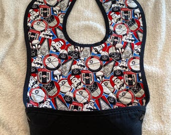 May include: A baby bib with a black bottom and a patterned top featuring Star Wars characters and logos in red, white, and blue. The bib has a black trim and snap closures.
