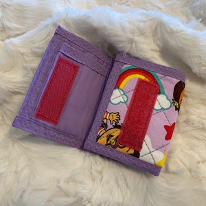 May include: A purple fabric wallet with a rainbow and cartoon character design. The wallet has a velcro closure and a pink fabric strap.
