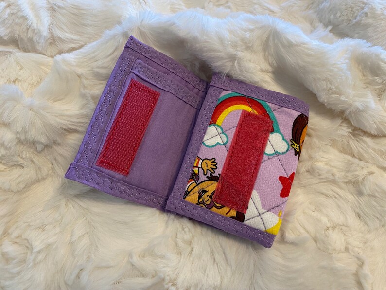 May include: A purple fabric wallet with a rainbow and cartoon character design. The wallet has a velcro closure and a pink fabric strap.